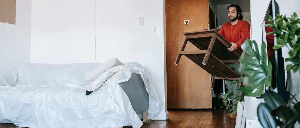 A man carrying a timber table through an apartment doorway past a sofa wrapped in plastic