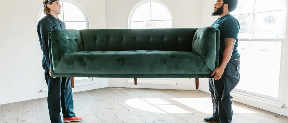 Two men carrying a green sofa across a bright, empty room with arched windows