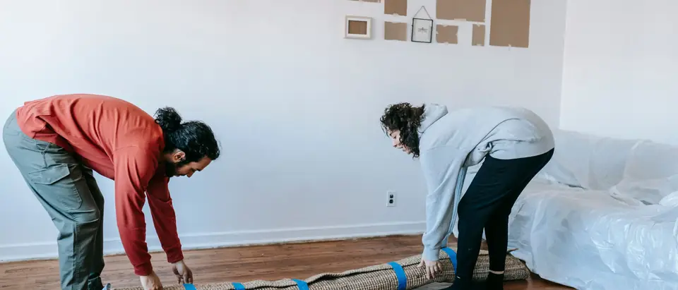 Two people rolling up a rug in a living room with the sofa wrapped in plastic